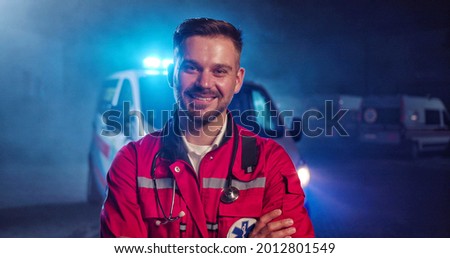 Portrait of Caucasian happy young male paramedic in red uniform smiling to camera and standing outdoor. Ambulance car on background. Attractive cherful male doctor at night shift. Call 911