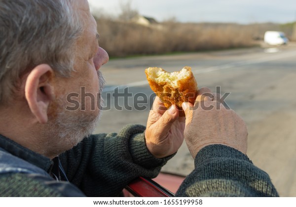 Portrait of Caucasian hungry senior\
driver eating patty leaning his elbows on the car\
door