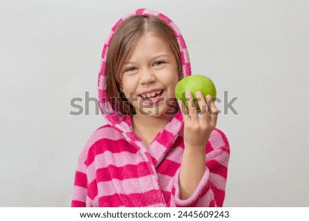Portrait of caucasian little girl in bathrobe with hood on head holding green apple looking at camera on white background