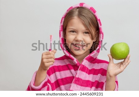 Portrait of caucasian little girl with open wide smile in bathrobe with hood on head holding toothbrush and green apple looking at camera on white background