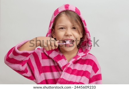 Portrait of caucasian little girl with open wide smile in bathrobe holding toothbrush close to teeth looking at camera on white background