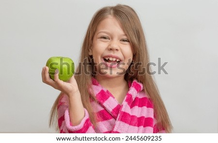 Portrait of caucasian little girl with open wide smile in bathrobe holding green apple in hand looking at camera on white background