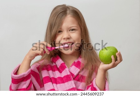 Portrait of caucasian little girl with open wide smile in bathrobe holding green apple and toothbrush looking at camera on white background