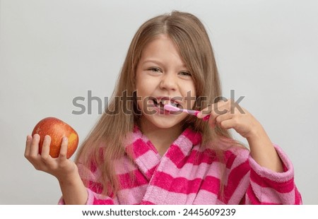 Portrait of caucasian little girl with open wide smile in bathrobe holding red apple and toothbrush looking at camera on white background