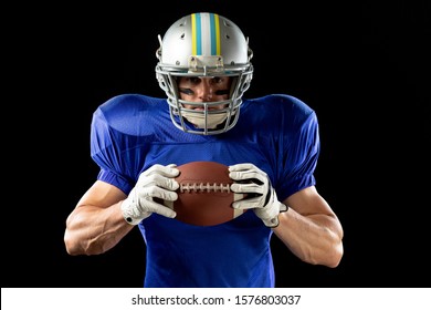 Portrait Of A Caucasian Male American Football Player Wearing A Team Uniform, Pads And A Helmet, Stripes Of Eye Black Under His Eyes, Holding A Football In Two Hands