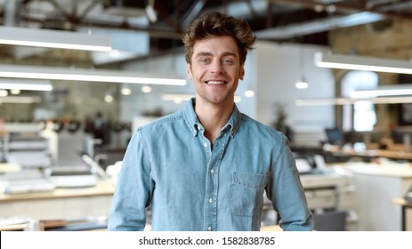 Portrait Of Caucasian Male Worker Wearing Denim Shirt Looking At Camera With A Smile While Standing In Co-working Space. Horizontal Shot. Front View. Selective Focus