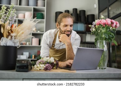 Portrait Of Caucasian Man Entrepreneur Sitting In Own Flower Shop, Typing On Laptop. Young Male Employee In Floral Store Working On Computer. Florist Concept.