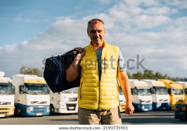 Portrait of caucasian\
mature man with bag on some-truck vehicles parking background.\
Truck driver worker 
