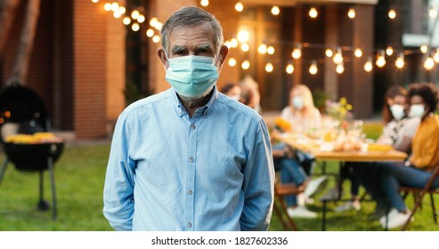 Portrait Of Caucasian Old Man In Medical Mask Standing At Yard And Looking At Camera. Senior Gray-haired Grandfather At Barbeque. Mixed-races Family Sitting At Dinner Table On Background. Outdoors.