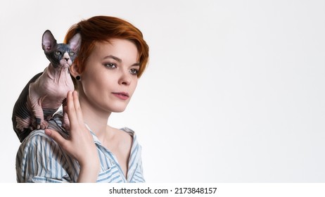 Portrait Of Caucasian Redhead Young Woman With Sphynx Cat Sitting On Her Shoulder. Female Hipster In Striped White And Blue Shirt. Studio Shot On White Background, Copy Space On Right. Part Of Series.