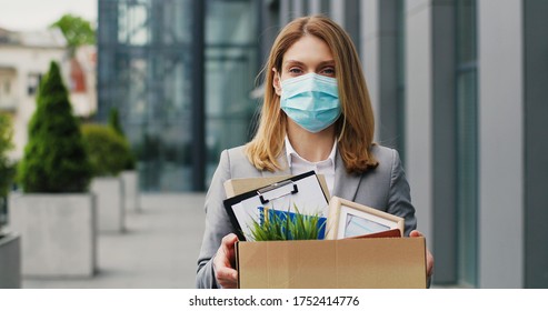 Portrait of Caucasian sad businesswoman in medical mask standing outdoor with box of stuff. Leaving business. Female office worker lost her job.