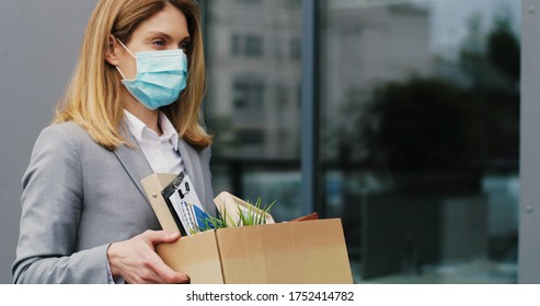 Portrait Of Caucasian Sad Businesswoman In Medical Mask Standing Outdoor With Box Of Stuff. Leaving Business. Female Office Worker Lost Her Job.