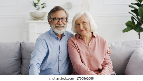 Portrait Of Caucasian Senior Couple Of Pensioners Sitting On Couch At Home. Old Man And Woman Looking At Camera On Sofa In Hugs In Living Room. Male And Female On Retirement.