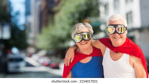 Portrait Of Caucasian Senior Couple Wearing Red Capes And Masks Standing Against Buildings In City. Love, Togetherness, Superhero, Power, Fantasy, Unaltered, Senior Citizens, Retirement, Awareness.