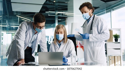 Portrait of Caucasian senior female doctor in medical mask sitting in hospital, typing on laptop computer and speaking with male healthcare colleagues at work. Coworkers in clinic during covid 19 - Powered by Shutterstock