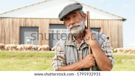 Portrait of Caucasian senior handsome man shepherd in hat standing outdoors, leaning on stick, looking down and rising face to camera. Old male farmer at his land. Close up.