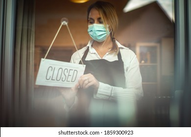 Portrait of a Caucasian woman with a black apron, wearing a face mask, standing behind a glass door putting the sign ''closed''