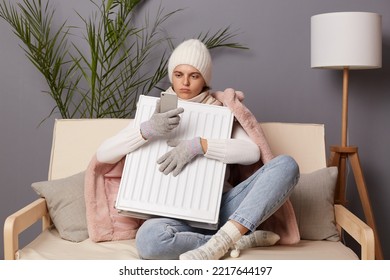 Portrait Of Caucasian Woman In Coat And Hat Sit In Cold Living Room Embracing Electric Heating To Get Warm And Using Cell Phone, Call To Service, Broken Central Heating.