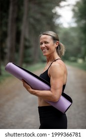 Portrait Of A Caucasian Woman In Her 40s Holding A Purple Yoga Mat Outside. She Is Smiling And Has Her Blonde Hair In A Ponytail. There Are Trees In The Background And She Is On A Gravel Road. 
