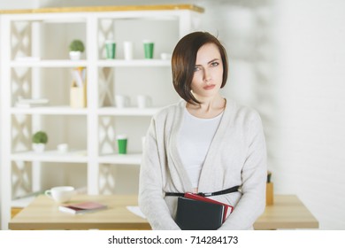 Portrait Of Caucasian Woman Holding Closed Book In Modern Office Interior. Knowledge And Information Concept 