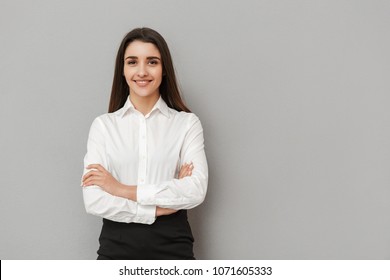 Portrait of caucasian woman with long brown hair in business wear smiling at camera and keeping arms folded isolated over gray background