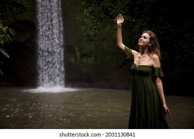 Portrait of Caucasian woman near the waterfall. Hand raised up. Enjoy nature. Water splash. Woman wearing green dress. Travel to Asia. Copy space. Tibumana waterfall, Bali, Indonesia