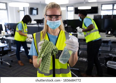 Portrait Of Caucasian Woman Wearing Hi Vis Vest, Gloves, Safety Glasses And Face Mask, Sanitizing Office With Colleagues In The Background. Hygiene In Workplace During Coronavirus Covid 19 Pandemic.