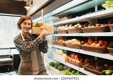 Portrait of Caucasian young adult employee is holding the basket of fruit and looking at camera with smile face. Cheerful saleswoman is working on stocking fruit on the shelves of the grocery store. - Powered by Shutterstock