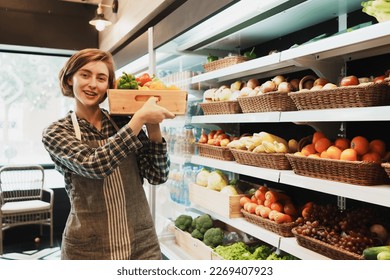 Portrait of Caucasian young adult employee is holding the basket of fruit and looking at camera with smile face. Cheerful saleswoman is working on stocking fruit on the shelves of the grocery store. - Powered by Shutterstock