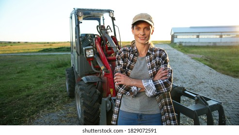 Portrait of Caucasian young beautiful woman in hat standing outdoor in field at big tractor and smiling to camera. Nice pretty female agriculture worker outdoor. Agricultural concept.
