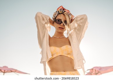 Portrait of caucasian young blonde with tied hair headscarf on background of clear sky. Girl holds her hands behind head, in sunglasses, yellow swimsuit, white shirt.