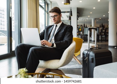 Portrait Of Caucasian Young Businessman Wearing Formal Black Suit Sitting On Armchair With Laptop Computer And Suitcase In Hotel Hall