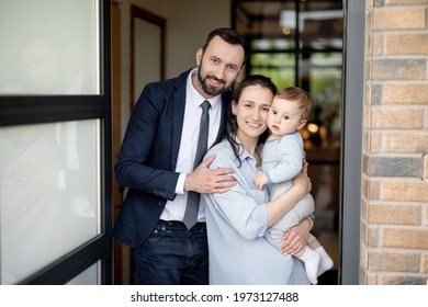 A Portrait Of Caucasian Young Family With Baby Standing In Front Of Their Front Door House Welcoming Guest At House. Happy Husband And Wife With One Year Old Baby Boy. Looking At Camera.