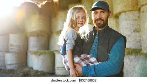 Portrait of Caucasian young handsome father holding on hands cute little daughter and smiling to camera at hay stocks in farm. Farming lifestyle. Countryside concept. Smiled man and small girl.