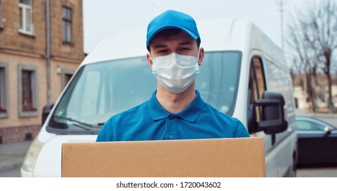 Portrait Of Caucasian Young Man Delivery Postal Worker In Gloves And Medical Mask Stepping In Front Of Camera With Parcel In Hands. Male Handing Carton, Delivery Bus Background.