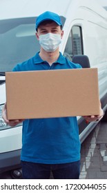 Portrait Of Caucasian Young Man Delivery Postal Worker In Gloves And Medical Mask Stepping In Front Of Camera With Parcel In Hands. Male Handing Carton, Delivery Bus Background.