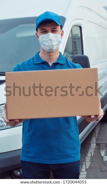 Portrait of Caucasian\
young man delivery postal worker in gloves and medical mask\
stepping in front of camera with parcel in hands. Male handing\
carton, delivery bus\
background.
