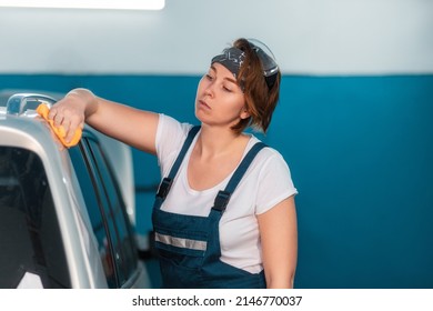 Portrait Of A Caucasian Young Woman In A Blue Coveralls Wiping A Car With A Rag. Car And Workshop In The Background. Work In An Auto Repair Shop And Garage.