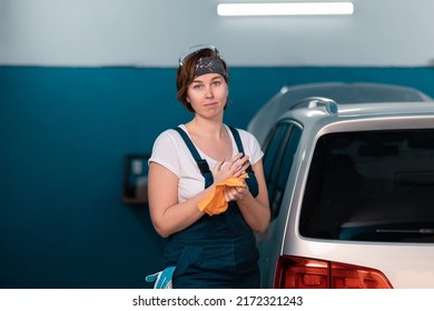 Portrait Of A Caucasian Young Woman In A Blue Coveralls Waping Her Hands With A Rag. Car And Workshop In The Background. Work In An Auto Repair Shop And Garage.