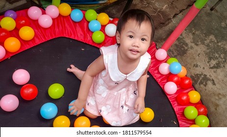 Portrait Of Charming 2 Years Old Cute Baby Asian Girl, Little Toddler Child With Adorable Short Black Hair Playing Trampoline With Colorful Small Ball At Home. Happy Kid On Vacation. Above View.