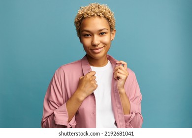 Portrait of charming adorable elegant smiling african american millennial woman with short blond curls touching collar of pink fashionable silk shirt isolated on blue studio background - Shutterstock ID 2192681041