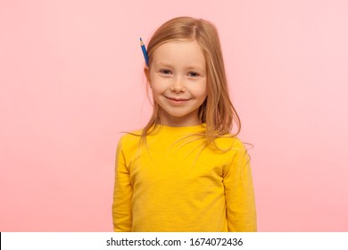 Portrait Of Charming Adorable Little Girl With Ginger Hair Holding Pencil Behind Ear And Smiling To Camera, Development Of Creative Child Abilities. Indoor Studio Shot Isolated On Pink Background