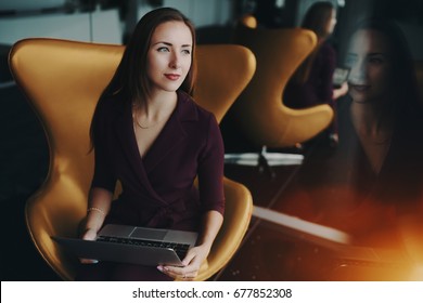 Portrait of charming adult caucasian businesswoman sitting on yellow elegant armchair with modern laptop next to mirror in luxury office with multiple reflections and wistfully looking aside