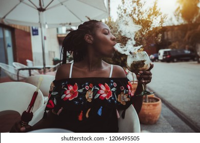 Portrait of a charming African girl sitting in a street cafe and making smoke in the glass of cocktail; young black female outdoors in a bar play with a hookah while drinking delicious mojito