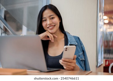 A portrait of a charming Asian woman in a denim jacket is working remotely from a coffee shop in the city, sitting at a table with her laptop and smartphone in her hand, smiling at the camera. - Powered by Shutterstock