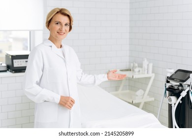 Portrait of charming beautician in white lab coat standing near daybed covered with sheet. She is looking at camera and smiling 