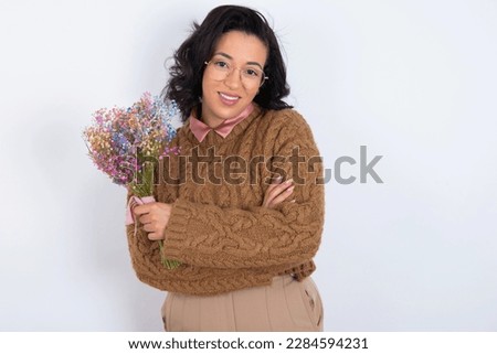 Portrait of charming beautiful woman holding a bouquet of flowers wearing knitted sweater over white background standing confidently smiling toothily with hands folded