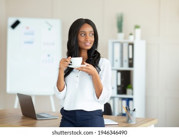 Portrait of charming black female manager in formal wear drinking coffee at contemporary office. Happy African American busineswoman taking break from work routine, enjoying hot drink