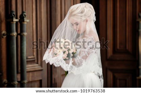 Portrait of a charming blonde bride in a white dress with lace, covered with veil, holding a wedding bouquet in her hands