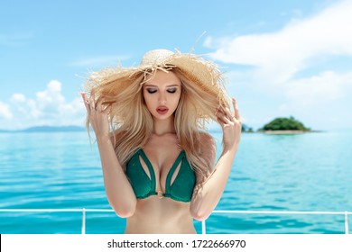 Portrait of a charming, charming blonde with bright makeup, in a straw hat, in a green bikini, posing, standing on the background of a lonely island. Endless blue sea. Closed eyes.